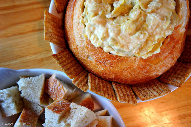 A close up of a display of a bread bowl with a dip inside of it, lined with crackers and next to a bowl of chopped bread.