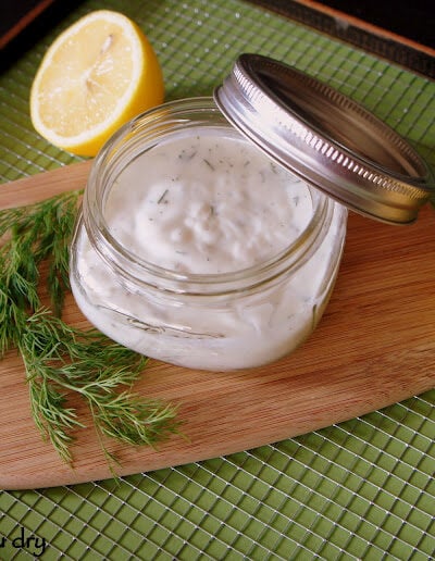 A small jar of Creamy Dill Dressing displayed on a table next to some fresh dill and a lemon half