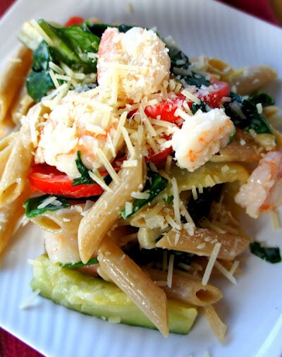 A close up of pasta displayed on a plate  topped with veggies, shrimp and cheese