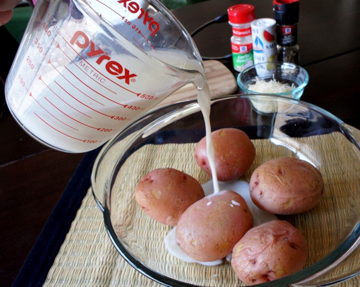 A glass bowl with 5 baked potatoes with buttermilk being poured into it