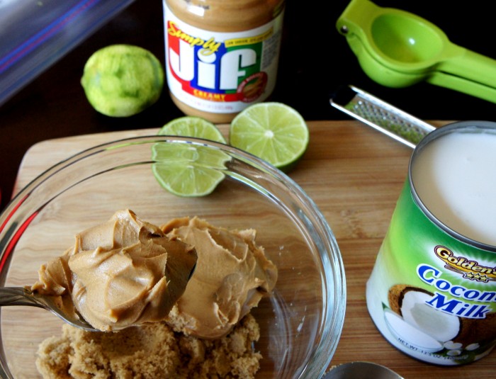 A bowl with dipping sauce ingredients on a cutting board with a side of a halved lime, a can of coconut milk & jar of PB