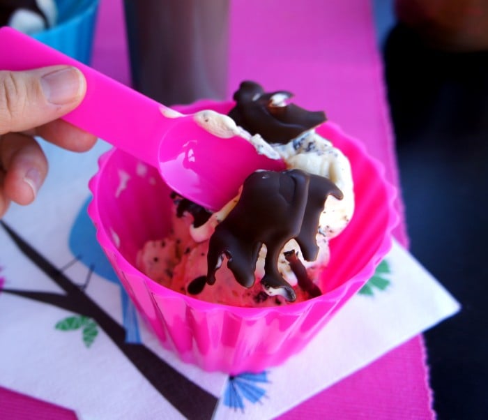 A spoon cracking the hardened chocolate sauce on top of a scoop of ice cream in a bowl