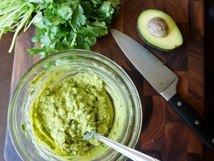 Blended avocado in a bowl sitting on a cutting board next to a knife, a half of avocado and some herbs