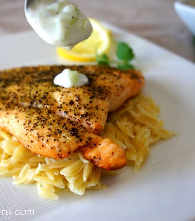 A spoon dripping Tzatziki Sauce on a Greek Salmon Fillet on a bed of noodles.