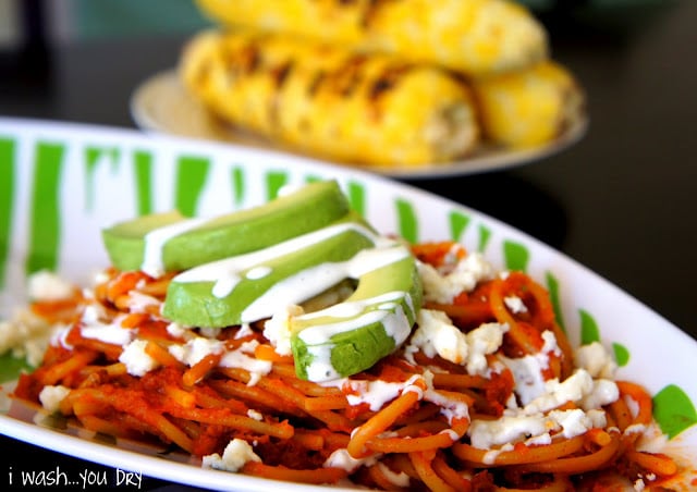 A bowl of red pasta with slices of avocado on top drizzled with a white sauce