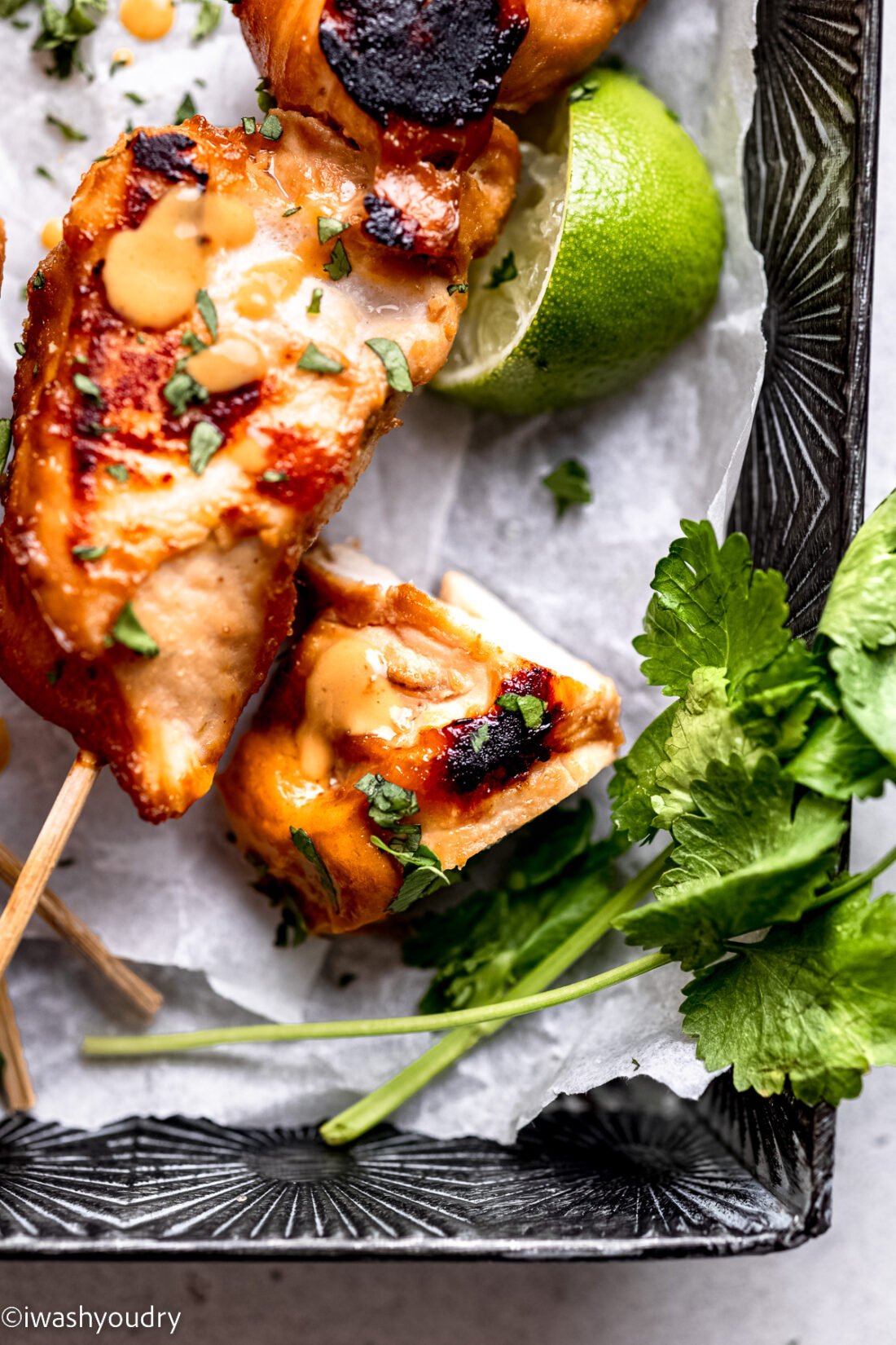 Piece of cooked Thai chicken satay in metal pan with cilantro and lime slice. 