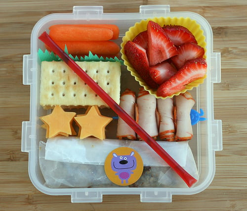 A bento lunch idea with carrot sticks, strawberry slices, crackers, star shaped cheese slices and rolled meet