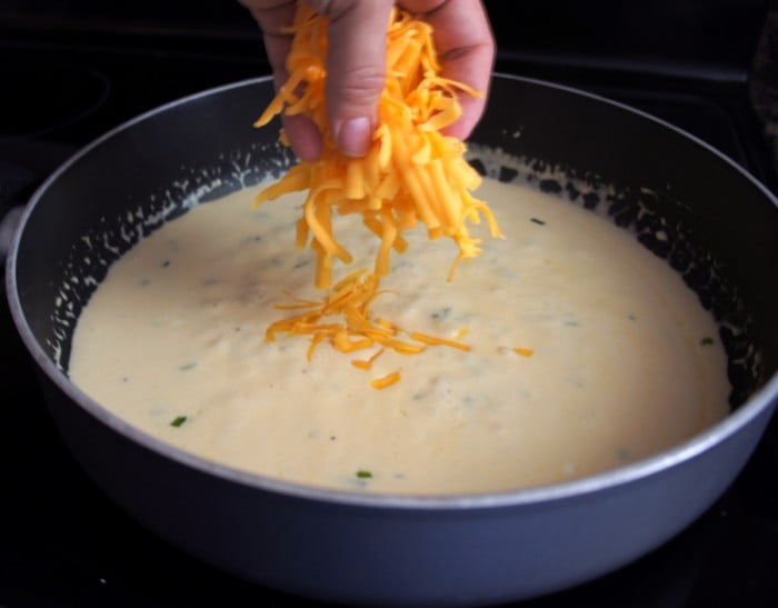 A hand adding a handful of orange shredded cheese to a pan of cream sauce