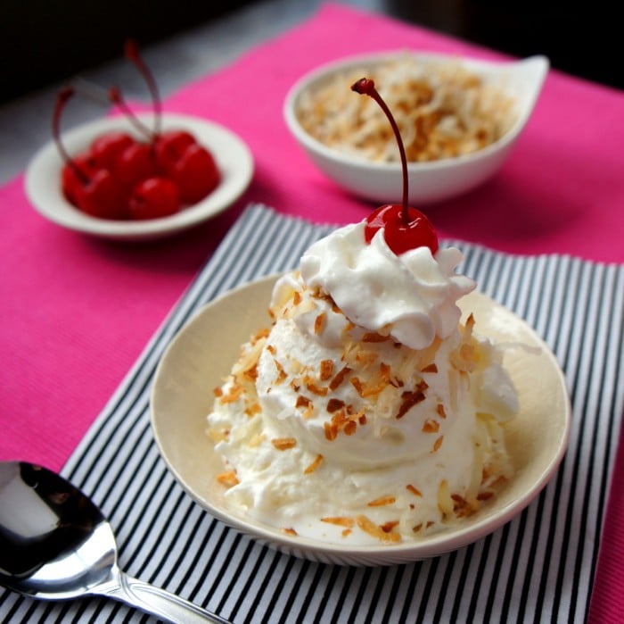 A bowl on a table with scoops of ice cream stacked onto of eachother, topped with nuts, whipped cream and a cherry
