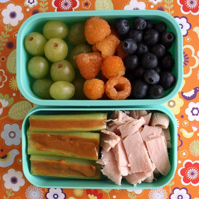 A lunch container with grapes, berries and blueberries and another container with celery sticks with peanut butter and slices of meat