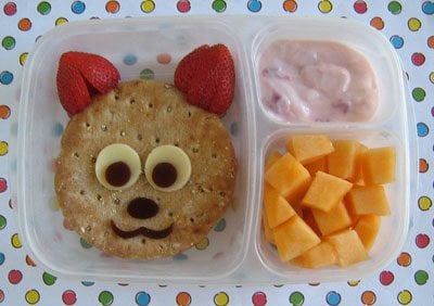 A bento box with a sandwich shaped cat using strawberries for ears, yogurt and cheese squares