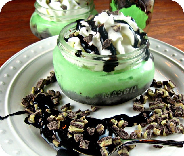  Mint Brownie Grasshopper Pie in a small mason jar topped with whipped cream and drizzled chocolate displayed on a plate
