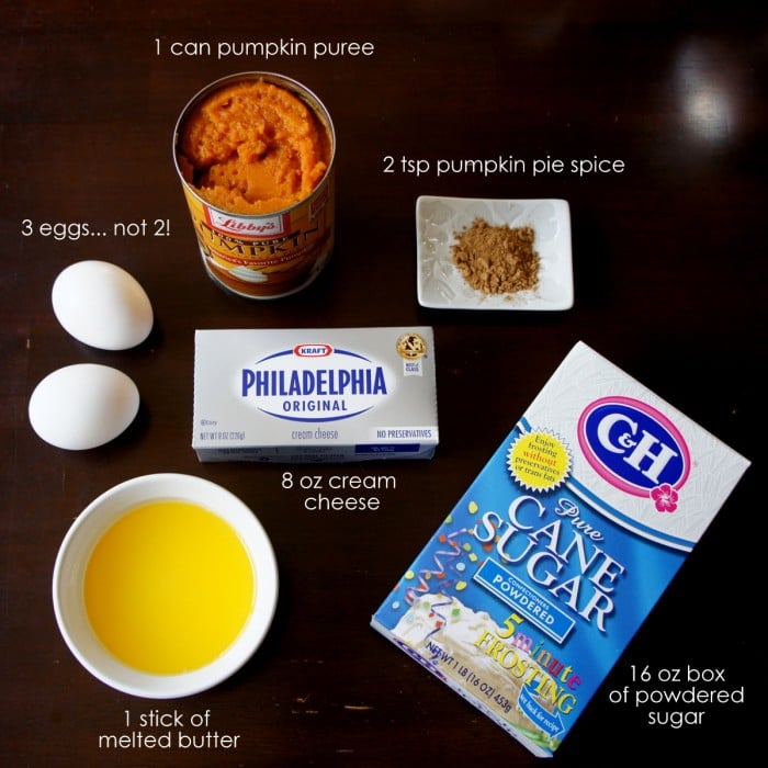 Ingredients for a recipe laid out on a table, with eggs, can of pumpkin puree, bar of cream cheese, pumpkin pie spice, melted butter and powdered sugar