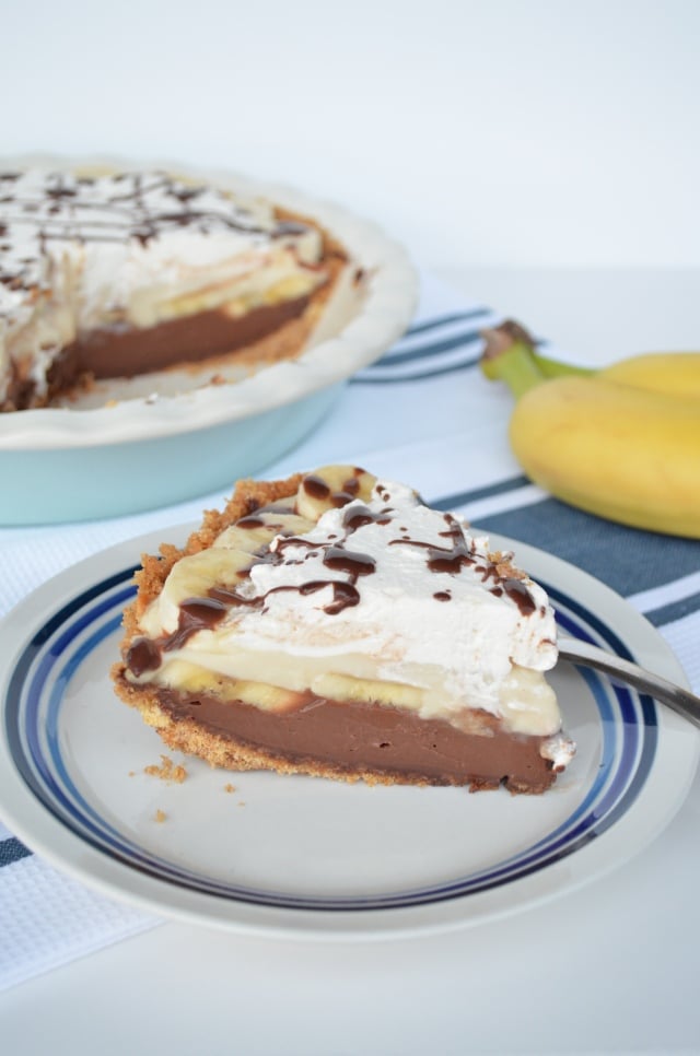 A slice of Black-Bottom Banana Cream Pie on a plate next to a couple of bananas and the full pie