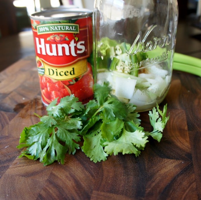 A mason jar with chopped onions in it on a table with a can of diced tomatoes and cilantro leaves next to it