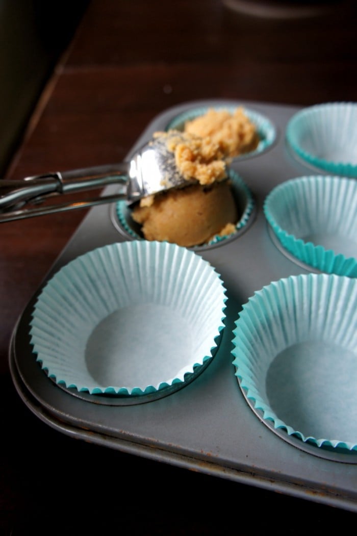 Muffin wrappers in a muffin tin with a scooper adding batter to one