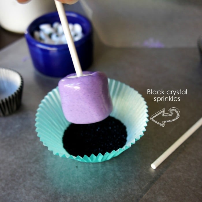 A purple dipped marshmallow pop being dipped in a cupcake liner filled with black sprinkles