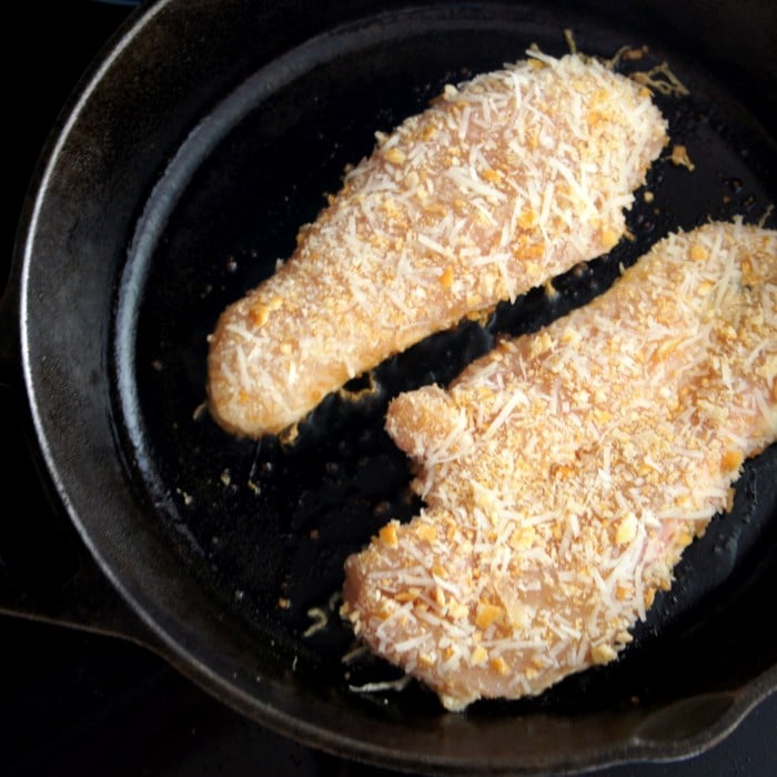 Two raw chicken breasts cooking in a pan with a breaded crust