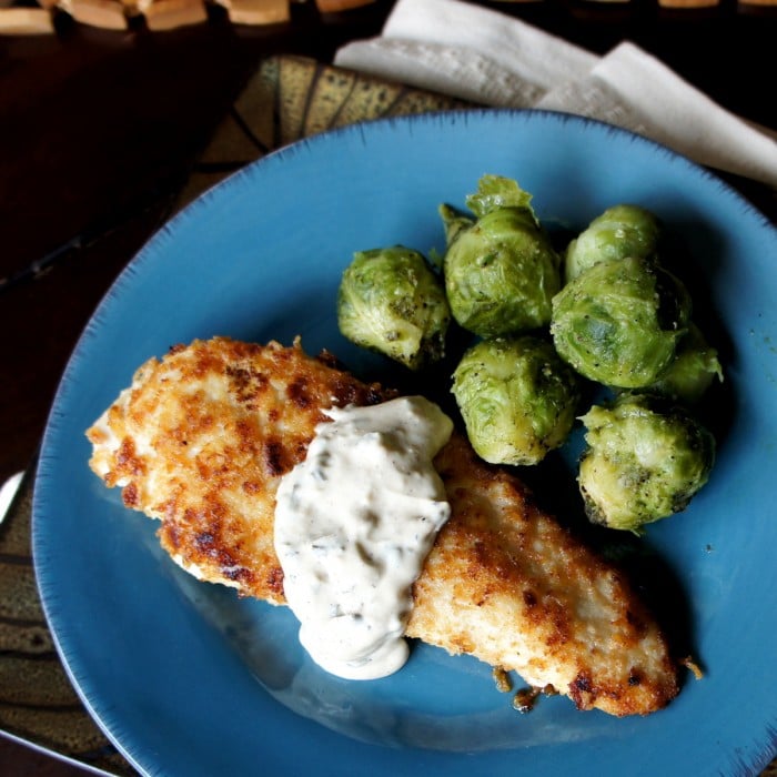 Cooked chicken breast on a blue plate topped with a white sauce and side of Brussels sprouts 
