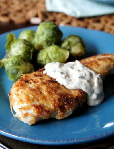 Cooked chicken breast on a blue plate topped with a white sauce and side of Brussels sprouts