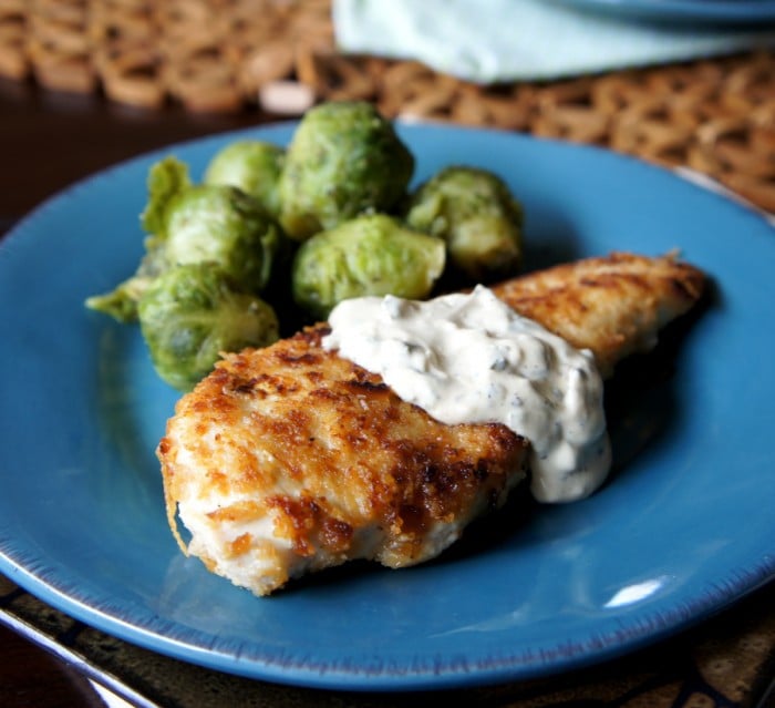 Cooked chicken breast on a blue plate topped with a white sauce and side of Brussels sprouts 