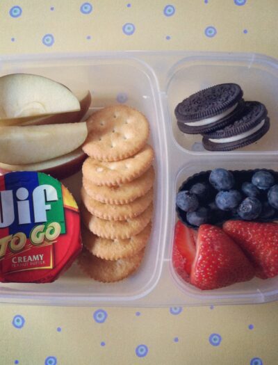 A Bento styled plastic container with dividers showcasing an easy lunch of crackers, peanut butter, apples, cookies and fruit