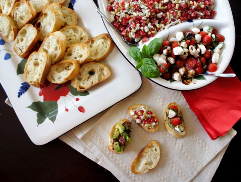 A close up of food on a table, with a tray of toasted baguette slices next to a tray of toppings and three Crostinis on a napkin