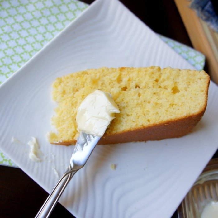 A piece of cornbread on a plate being spread with butter