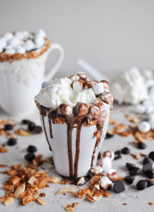 A min cup of overflowing hot chocolate with mini marshmallows topped with whipped cream and nuts