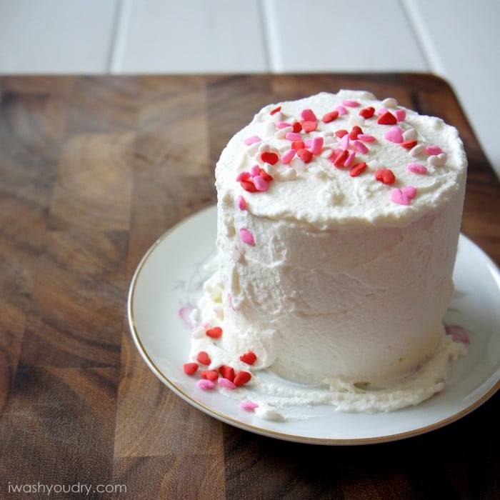 A white frosted cake on a plate with pink, red and white heart shaped sprinkles on top