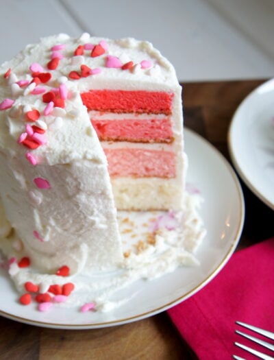 A 4 layer ombre cake with white frosting on a plate and a slice removed from it
