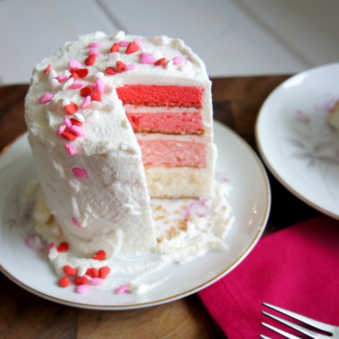 A 4 layer ombre cake with white frosting on a plate and a slice removed from it