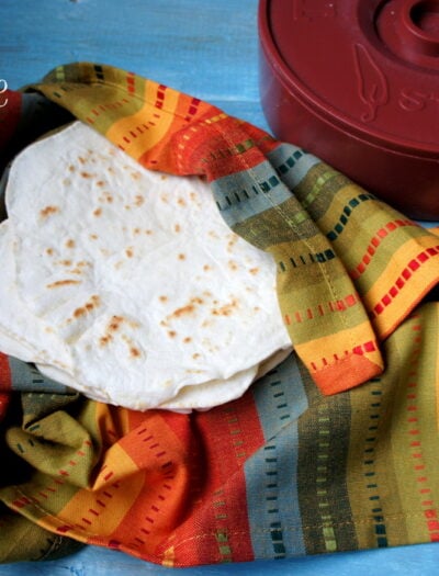 A pile of homemade tortillas in a colorful cloth next to a tortilla warmer