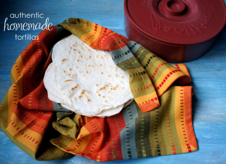 A pile of homemade tortillas in a colorful cloth next to a tortilla warmer