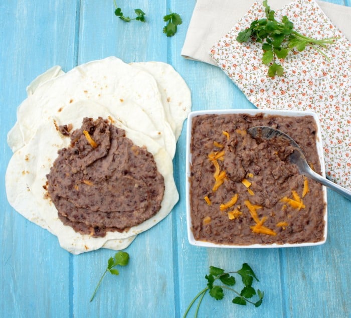 A square bowl of refried beans topped with cheese next to a small pile of tortillas with refried beans spread on one