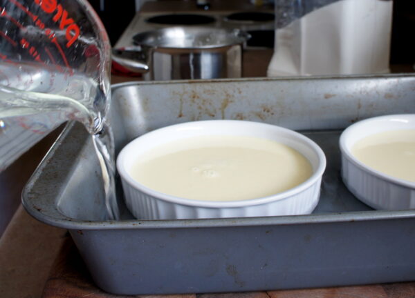 A ramekin filled with white liquid in a metal pan being filled with water