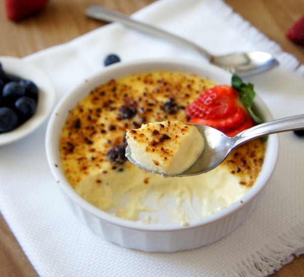 A spoon with creme brûlée on it over a ramekin of Blueberry Cheesecake Creme Brûlée with some of it already eaten