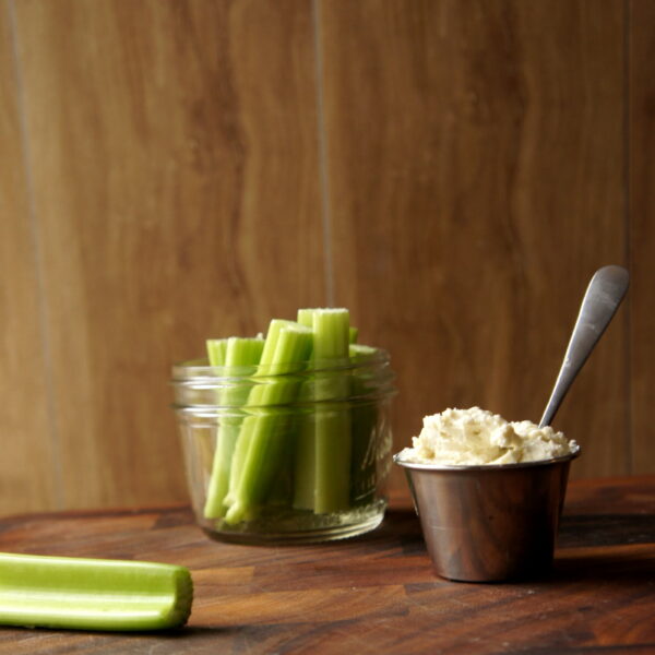 A short glass jar with celery sticks next to a small cup of dip