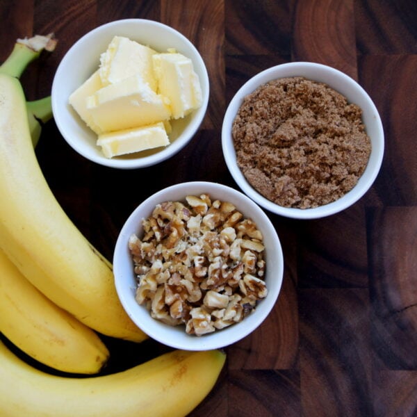 A wooden surface with a bowl of butter slices, a bowl of chopped walnuts, a bowl of brown sugar and 3 bananas next to them