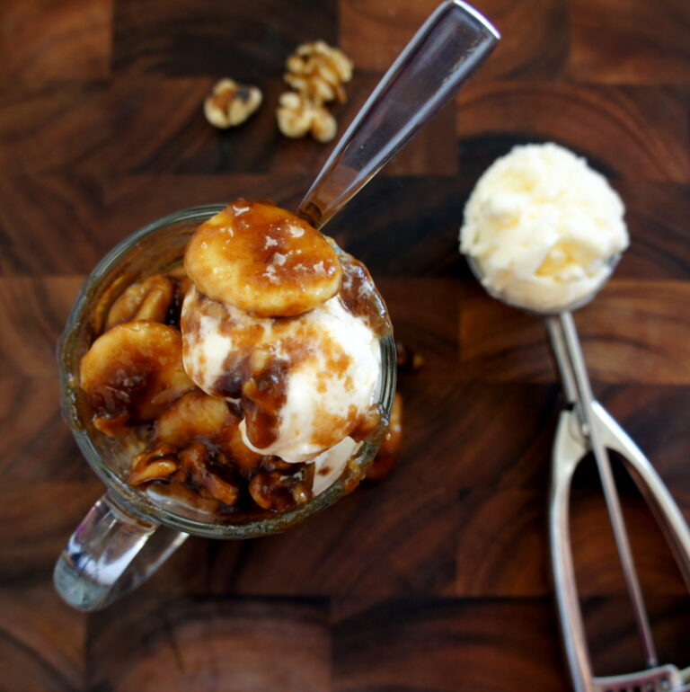 A look on from above of a glass mug filled vanilla ice cream topped with Banana Fosters and a ice cream scooper with vanilla ice-cream next to it