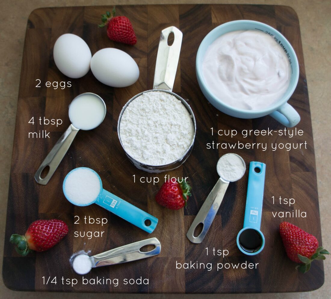 A group of ingredients on a wooden table with berries, includes eggs, flour, milk, strawberry yogurt, sugar, baking soda, baking powder, vanilla