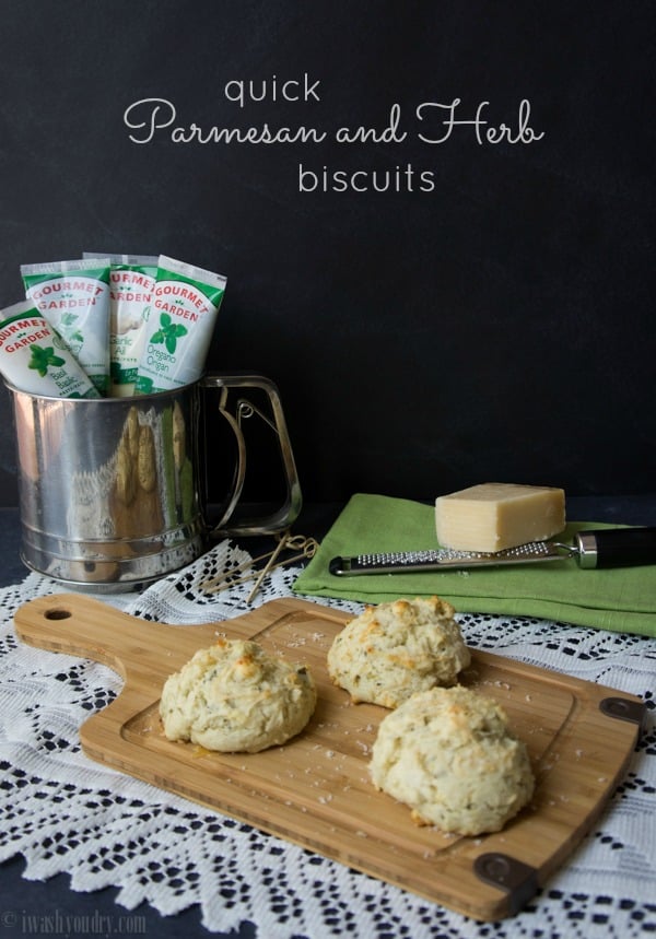 Three biscuits on a wooden cutting board surrounded by tubes of herbs and a block of cheese