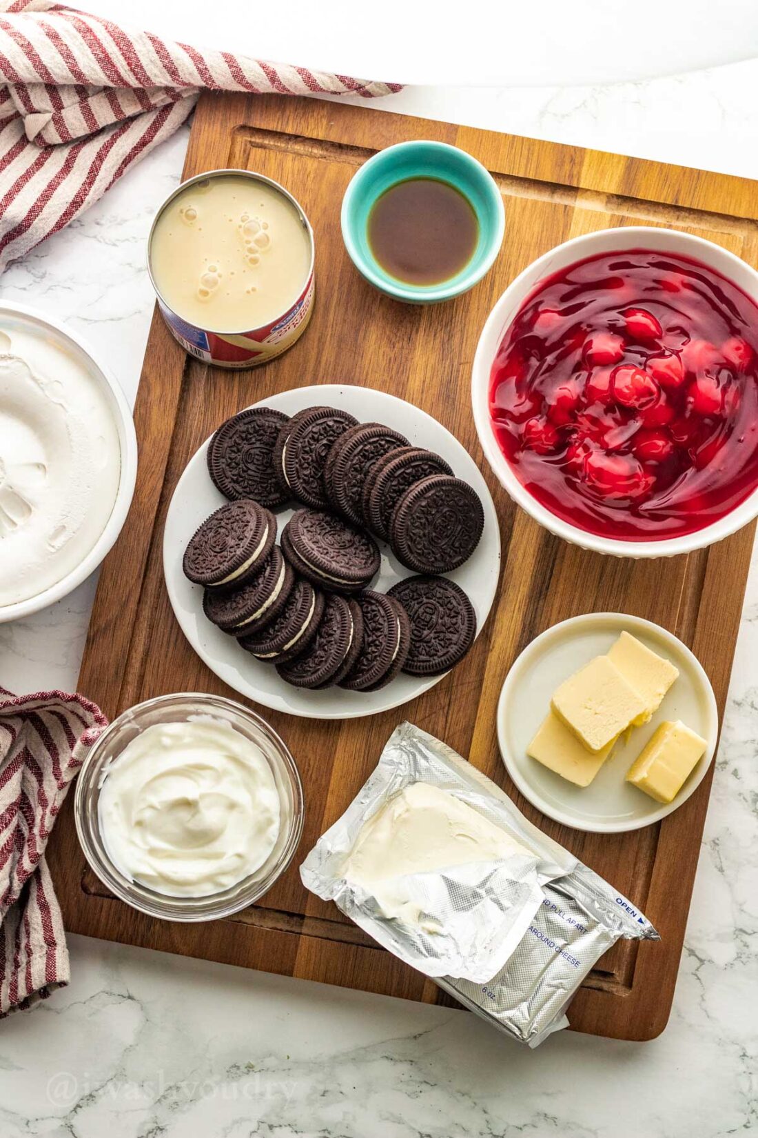 Oreos, cherries, cream cheese, and other ice cream ingredients on wood cutting board. 