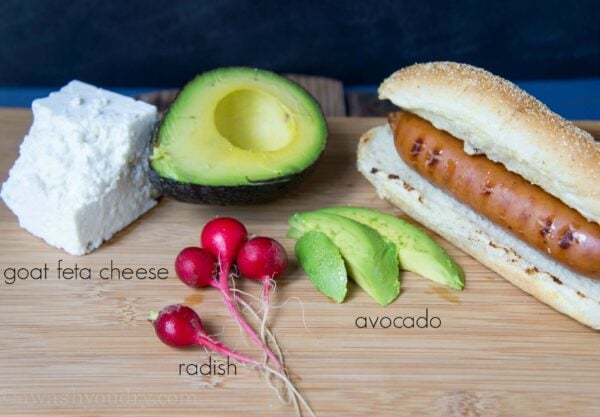 A wood surface with radishes, sliced avocado and a Polish dog in a bun