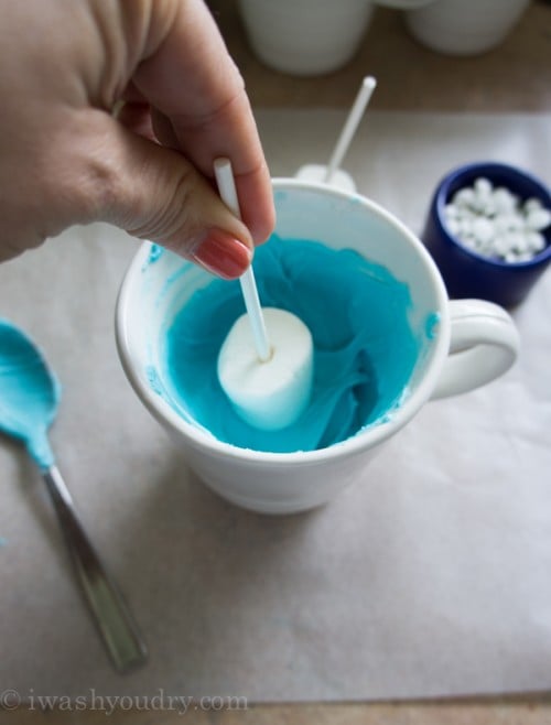 A marshmallow pop being dipped in a bowl of blue frosting