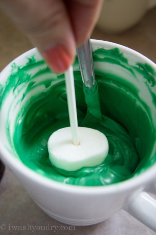 A marshmallow pop being rolled in a bowl of green frosting