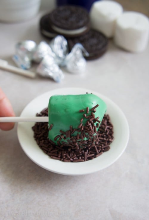A green frosted marshmallow pop being rolled in a small bowl of chocolate sprinkles