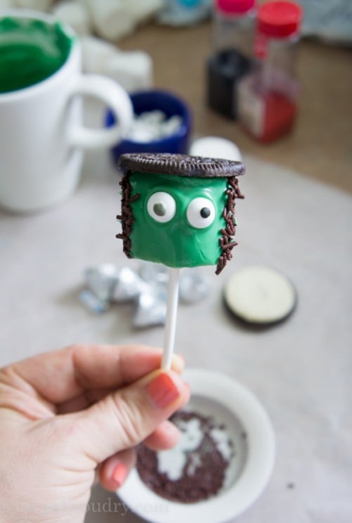 A close up of a green marshmallow pop with eyeballs and an Oreo cookie and chocolate sprinkles for hair