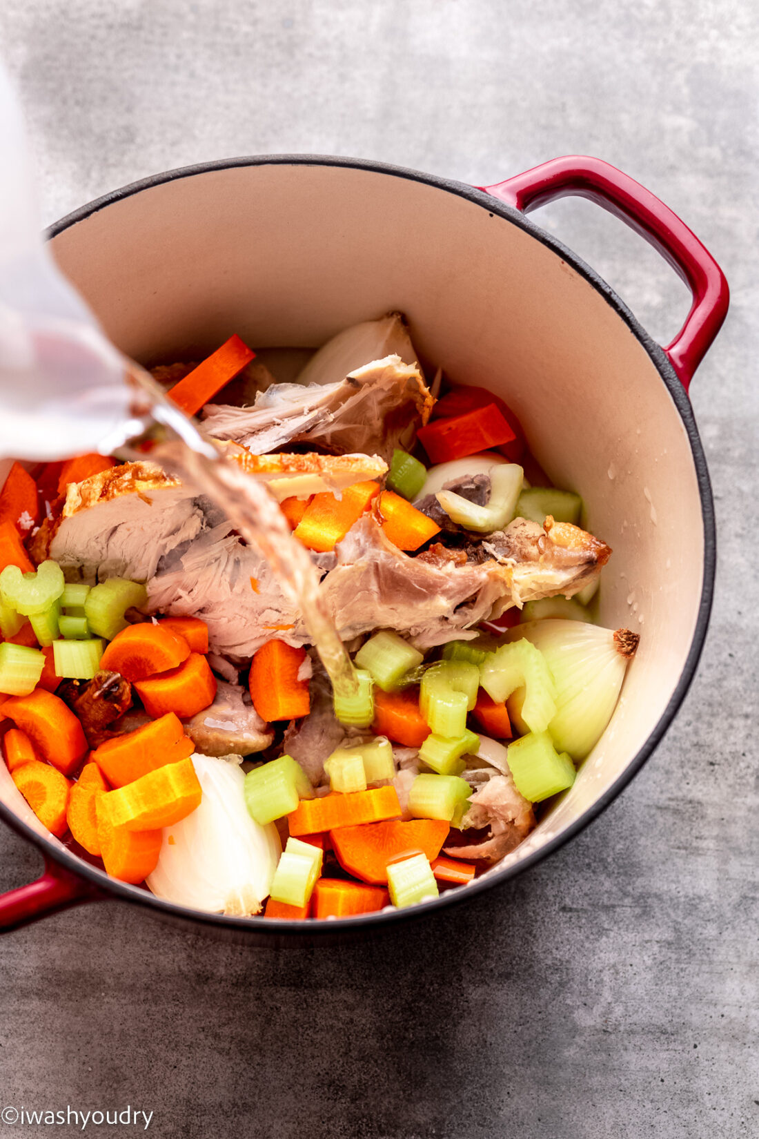 Carrots, celery, onion, and spices in red pot with water pouring. 