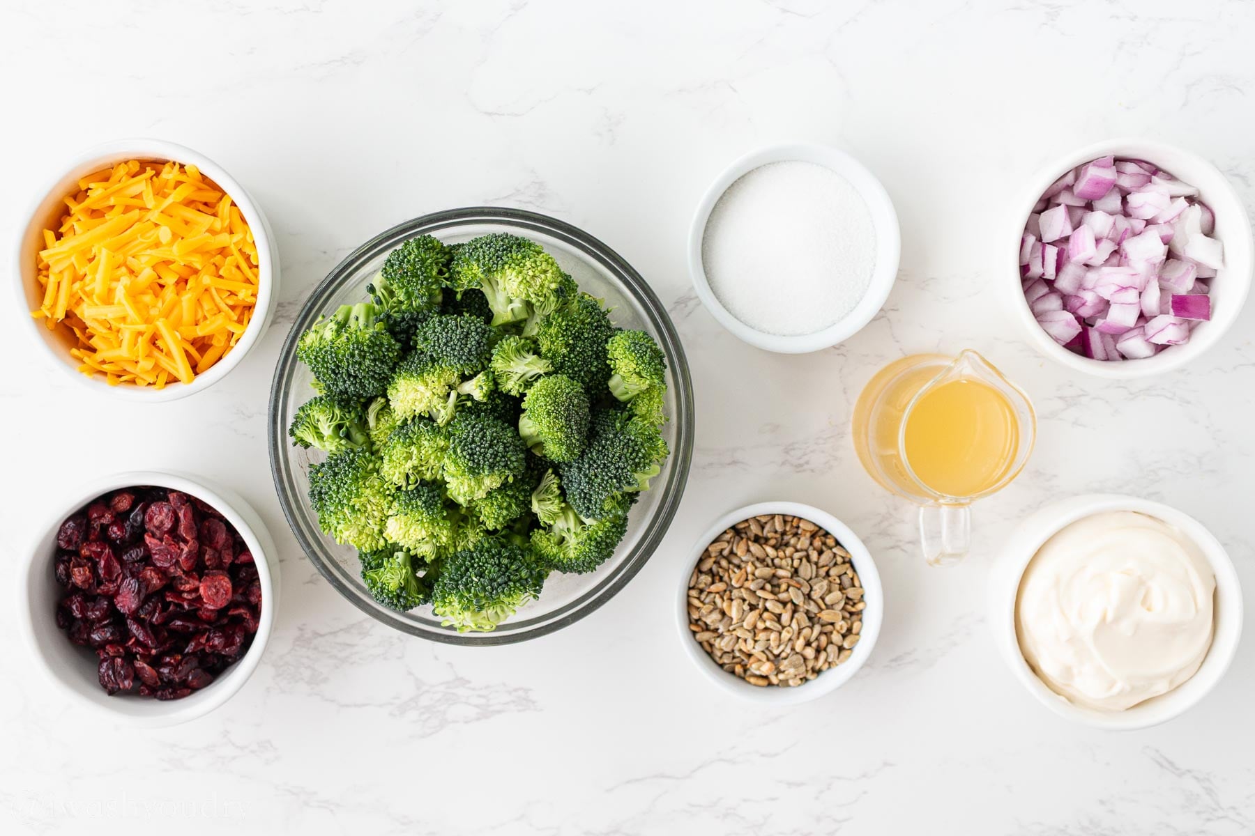Ingredients for broccoli salad on a white surface with cheddar cheese and sunflower seeds.
